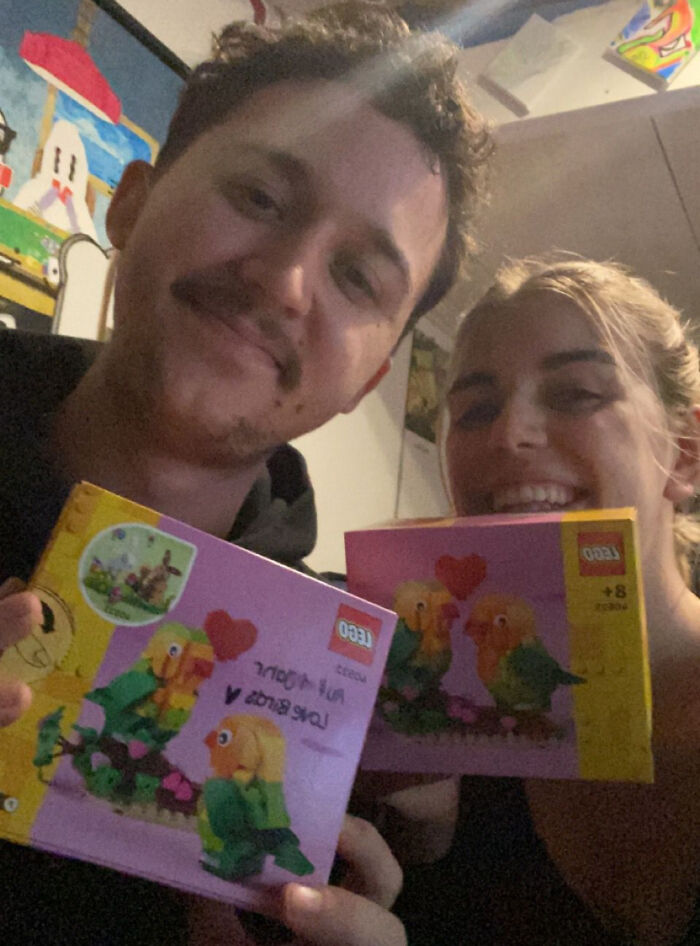 Couple smiling, holding lovebird Lego sets for a funny-wholesome-anniversary-surprise at home.