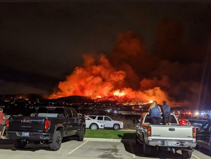 A large wildfire burns on a hillside, with people observing from trucks, highlighting nature disaster resilience.