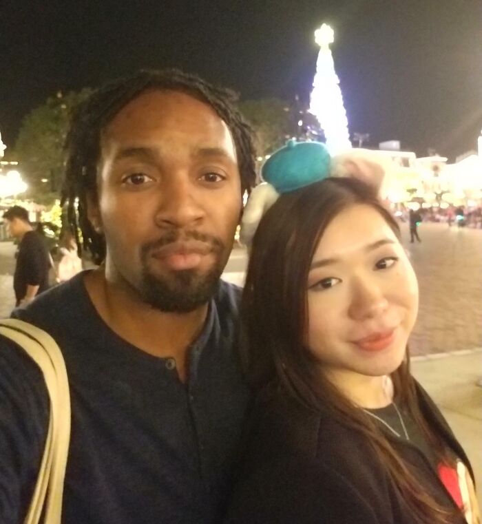 A couple smiling in front of a lit-up Christmas tree at night, showcasing long-distance love.