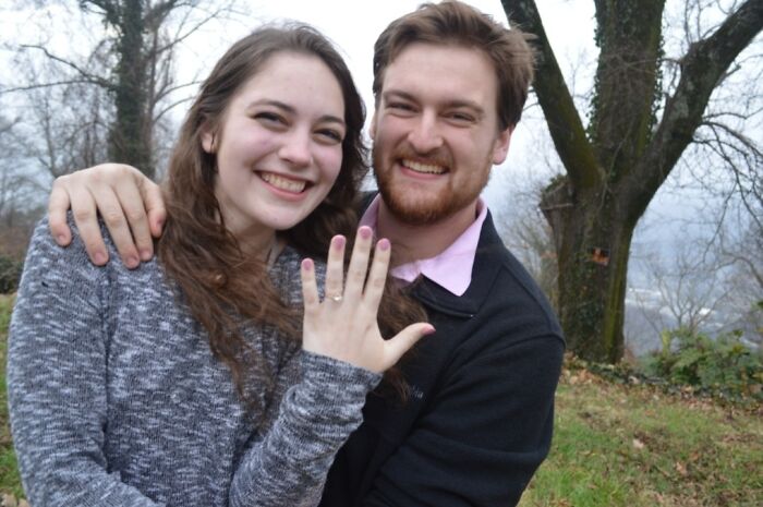 Couple smiling and showing an engagement ring, symbolizing that love always wins, outdoors with trees in the background.