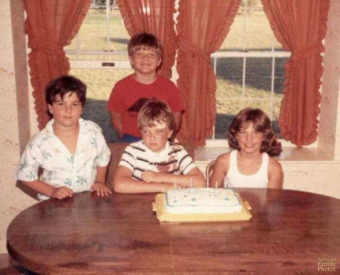 Four kids posing awkwardly around a birthday cake in a vintage family photo that feels both awkward and sweet.