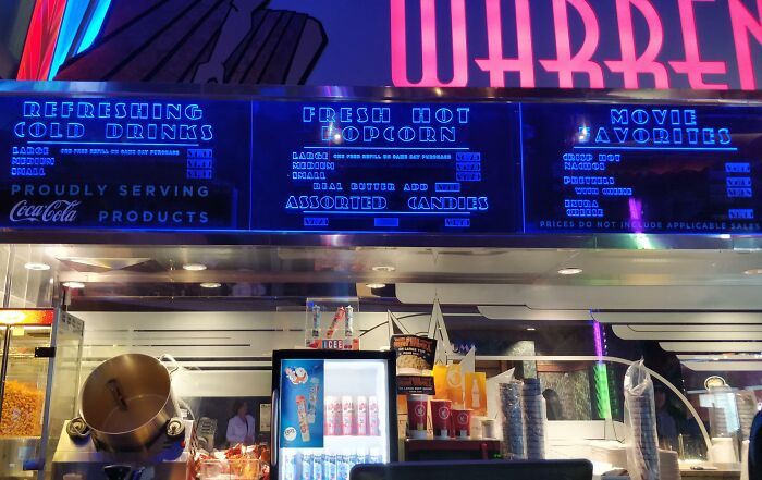 Unfortunate public interior design at a concession stand with difficult-to-read neon menu and cluttered counter space.