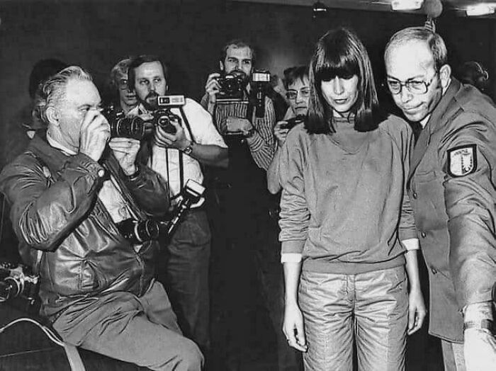 Black and white photo of a woman surrounded by photographers and a man in uniform, illustrating morally grey acts.