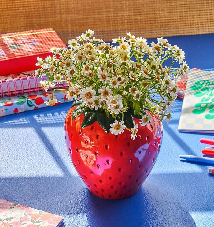 Strawberry-shaped vase with daisies, highlighting dopamine decor theme on a blue table with notebooks and pens.