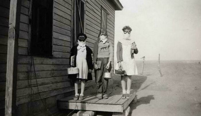 Three children wearing vintage protective masks and goggles standing on a wooden porch in a historical old image.