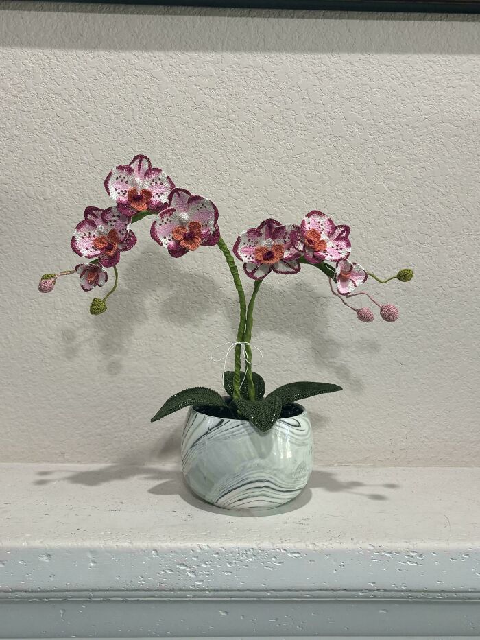 Crochet enthusiasts' detailed handmade pink orchid flowers with green leaves in a marble-patterned pot on a white ledge.