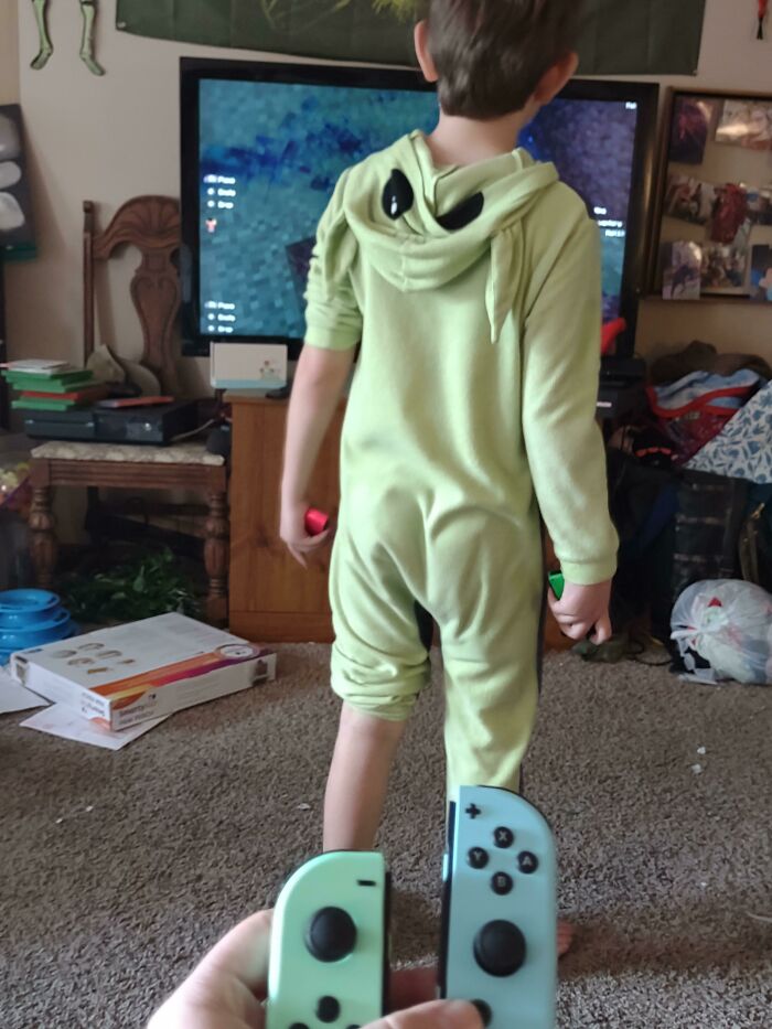 Young child in green onesie playing video game, with controllers visible in foreground.