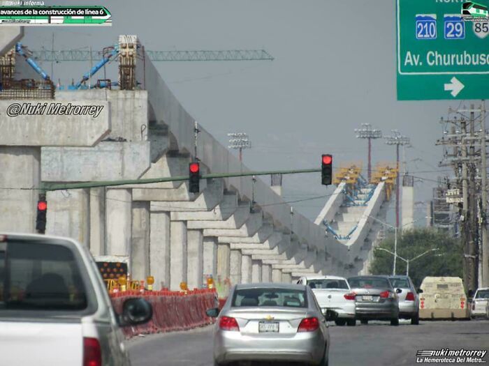 Construction of elevated concrete infrastructure for metro line 6 progressing alongside busy urban road with cars and traffic lights.