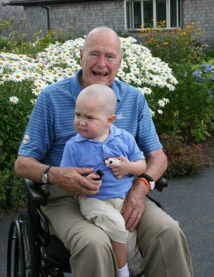 An elderly man and a young child, both smiling, sitting together in a garden of daisies; interesting and fascinating moment captured.