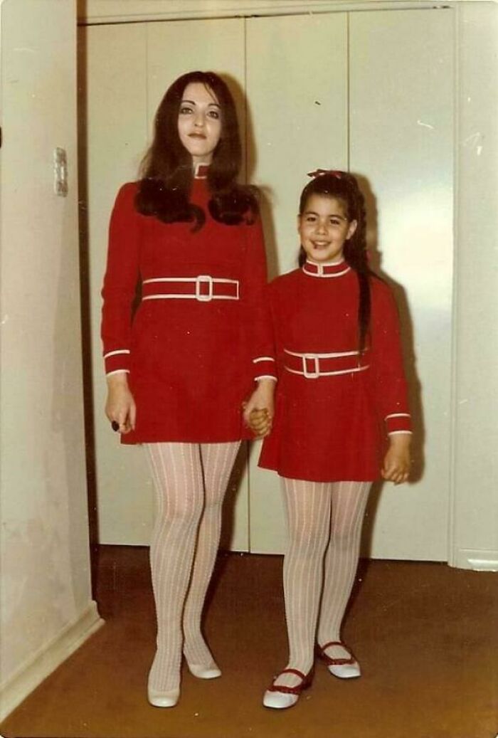 Two females in matching red vintage outfits and patterned tights holding hands in a hallway in a fascinating old image.