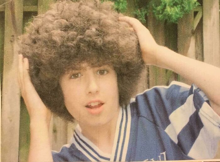 Young person with a large curly hairstyle, wearing a striped sports jersey, posing outdoors. Embarrassing blunder years moment.