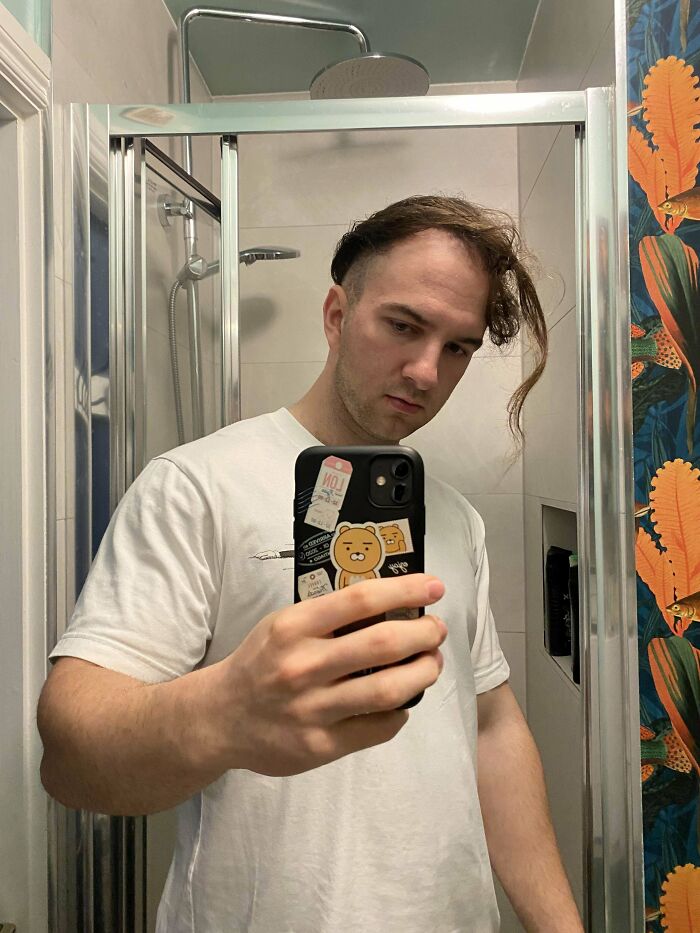 A man with an unfortunate haircut, long hair on one side and shaved on the other, looks in a mirror.