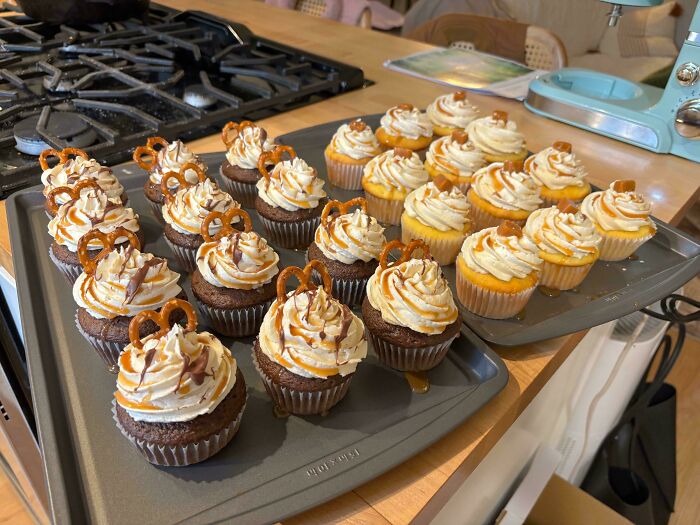 Culinary concoctions: pretzel-topped cupcakes with swirled frosting and caramel drizzle on baking trays in a kitchen.