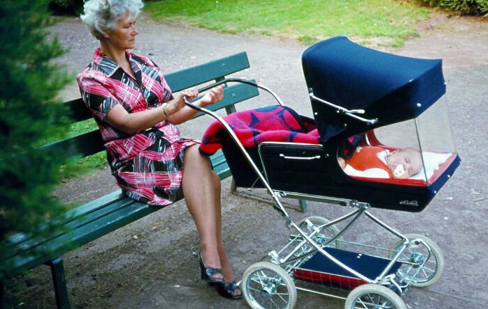 Woman sitting on a bench next to a vintage baby stroller with a clear protective cover in a park setting, old images.