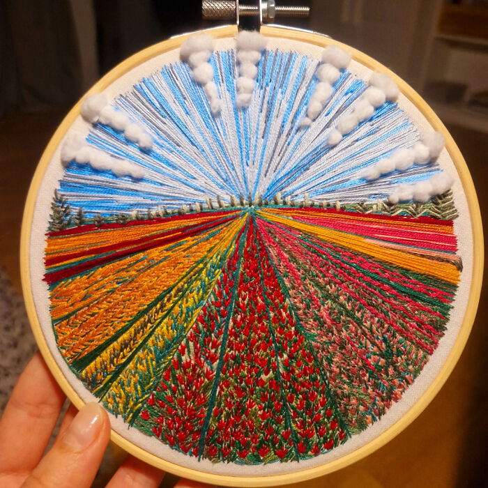 Hand holding embroidery hoop with colorful embroidery designs depicting fields, sky, and clouds in detailed stitching.