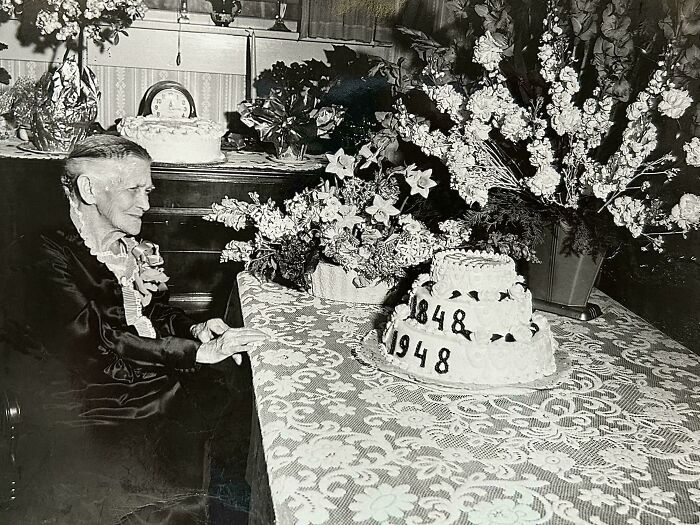 Elderly woman sitting by a decorated table with a 100th birthday cake from 1848 to 1948 in old images.