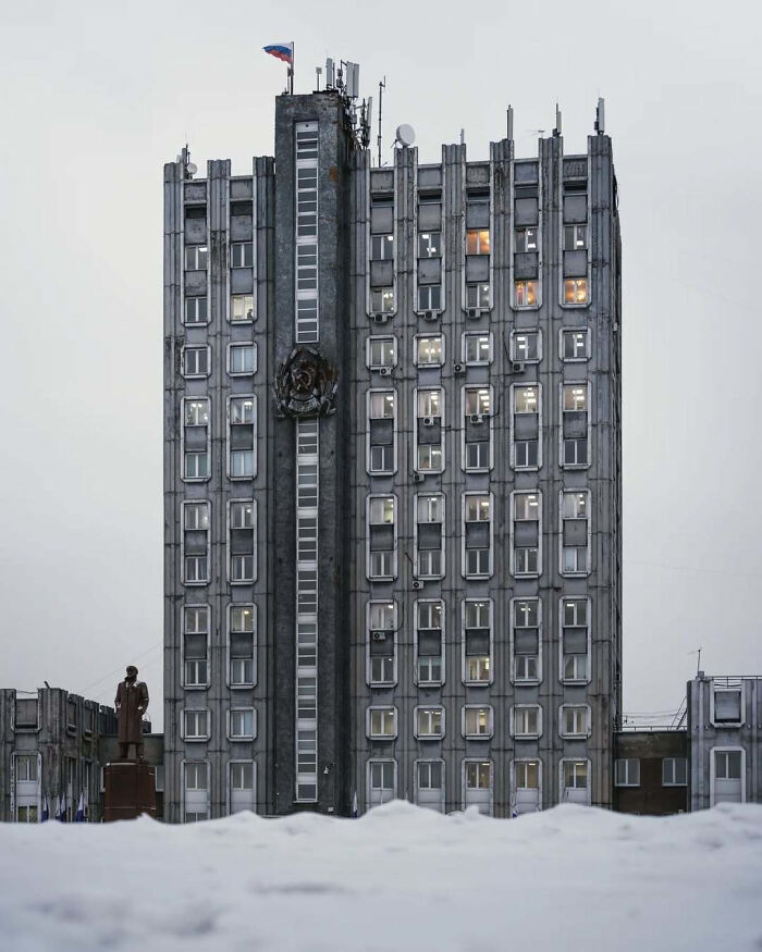 Eerie cold concrete building with many windows and a statue nearby, evoking supervillain headquarters vibes.