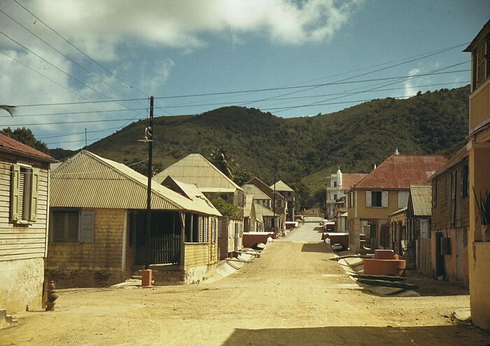 1940 color photo of a quiet street with vintage buildings in a small town, set against a backdrop of lush green hills.