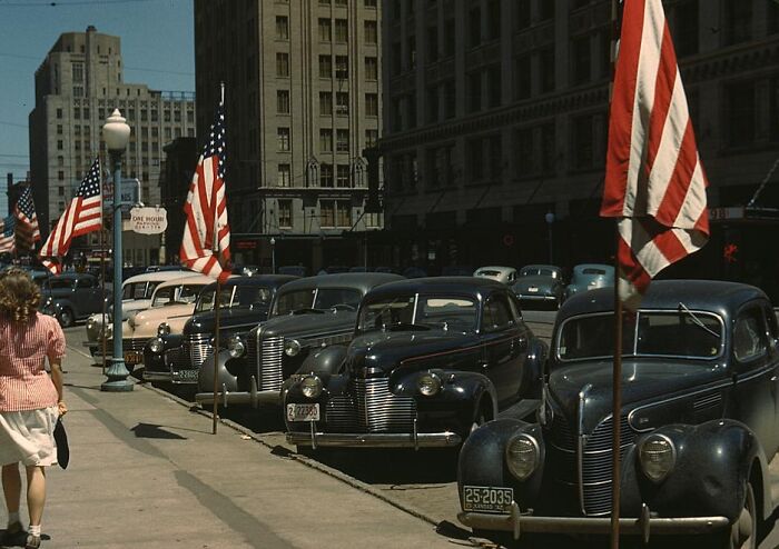 1940 color photo of vintage cars parked along a street with American flags and pedestrians in a city setting.