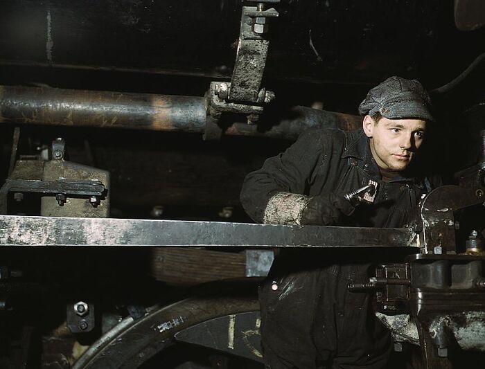 Young worker in a 1940 color photo, wearing a cap, focusing on machinery.