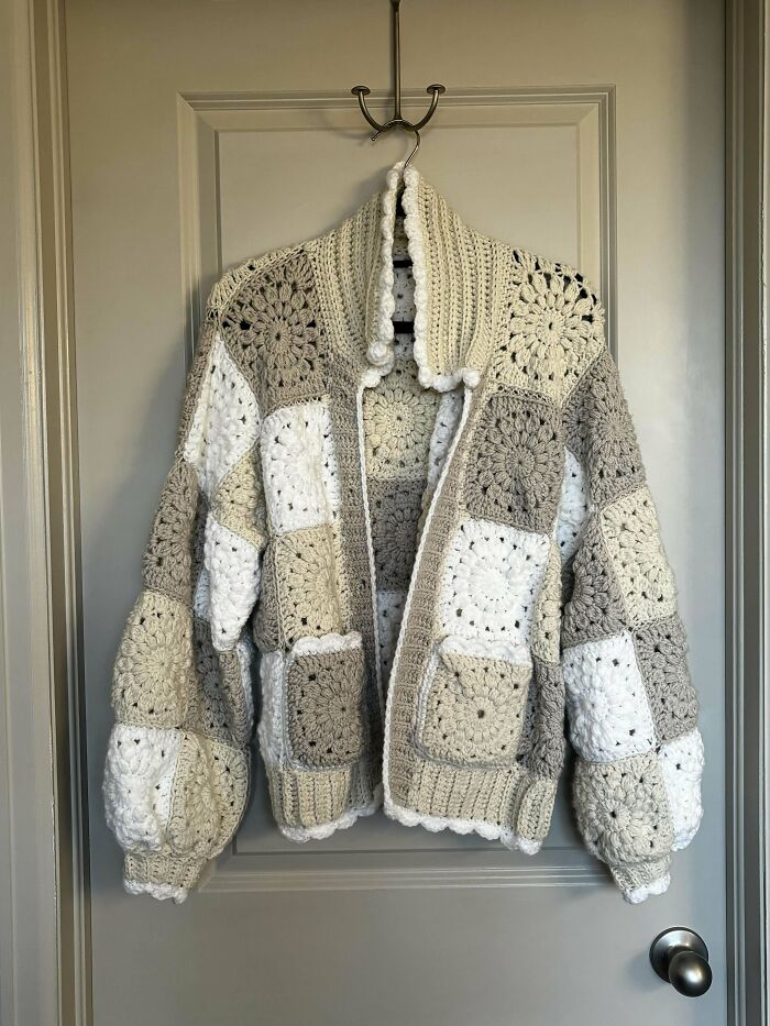 Chunky crochet cardigan made of beige and white granny squares hanging on a door, showcasing beautiful crochet masterworks.