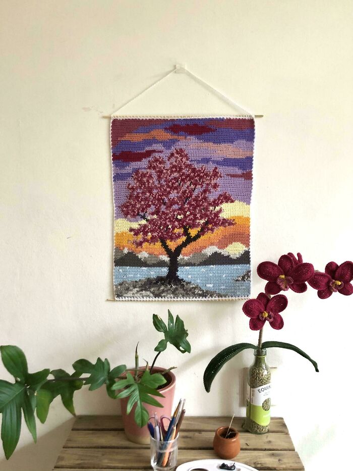Crochet wall hanging featuring a tree at sunset, displayed above a wooden table with plants and art supplies.