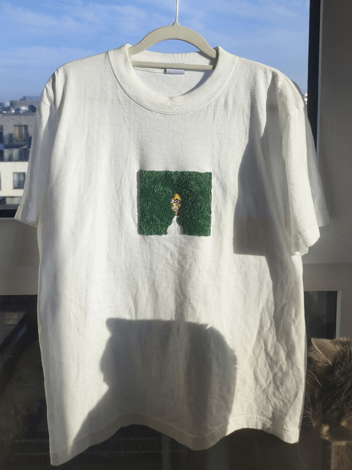 White t-shirt hanging on a hanger with an embroidered design of a character peeking through green foliage, showcasing embroidery skills.