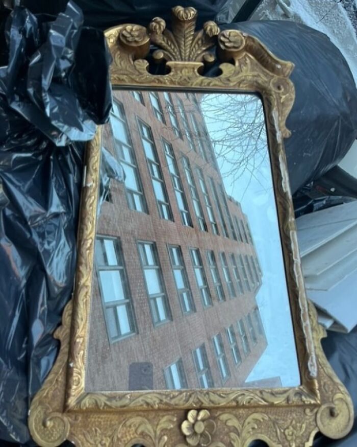 Ornate gold-framed mirror left on NYC curb, reflecting a building.