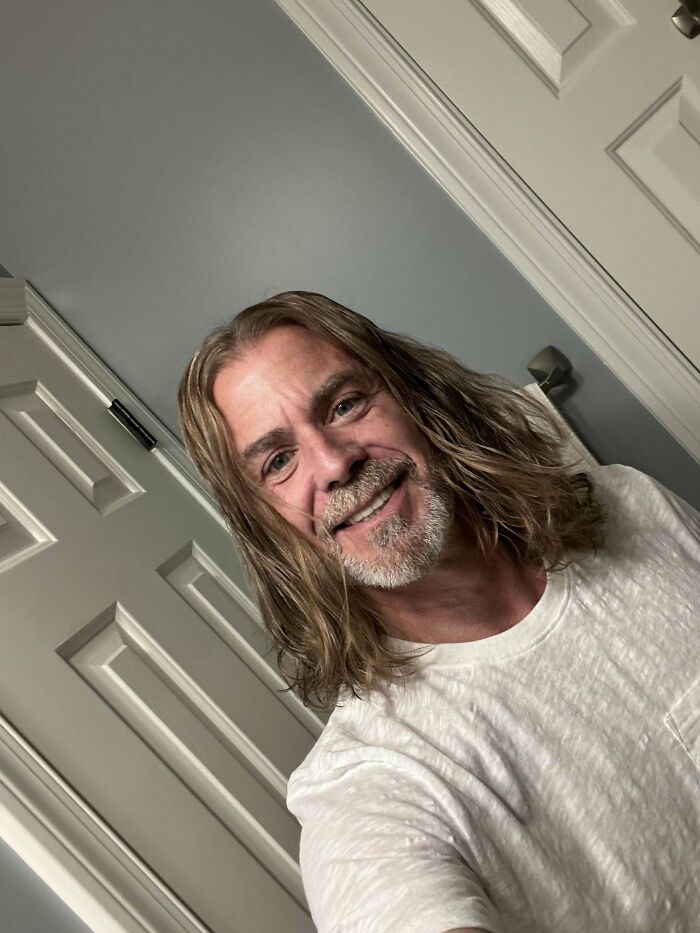 Man with fierceflow beautiful hair men style and a light beard smiling indoors wearing a white t-shirt.