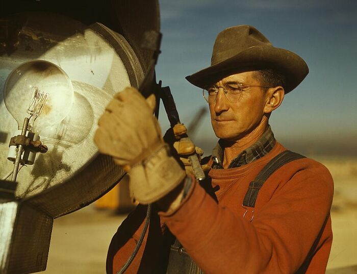 Man in 1940s attire, wearing glasses and a hat, adjusting a large vintage light fixture outdoors, in a color photo.