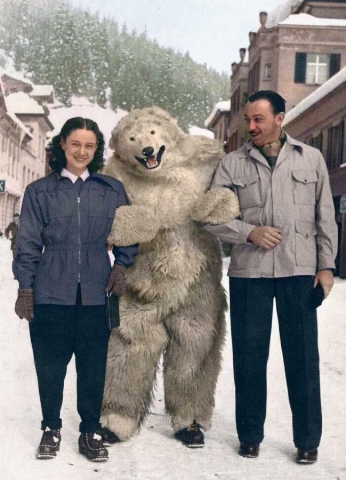 People in 1940 color photo posing with someone in a bear costume, set against a snowy street backdrop.