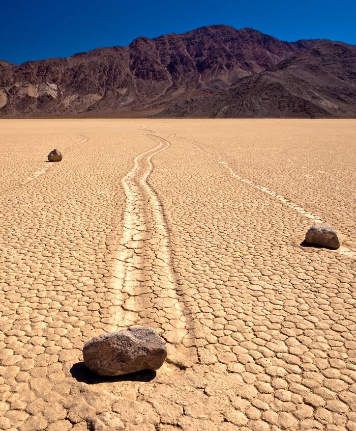 Rocks create tracks on sun-c*****d desert floor, showcasing strange natural phenomena under a clear blue sky.