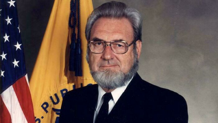 An older man with a beard, in a suit, standing in front of a U.S. flag and yellow banner, evoking childhood nostalgia.