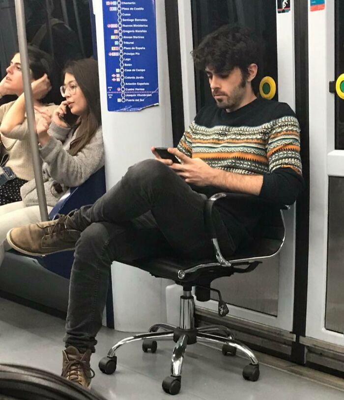 Man using a swivel chair as a seat on a subway train, browsing his phone, showcasing funny train passengers.