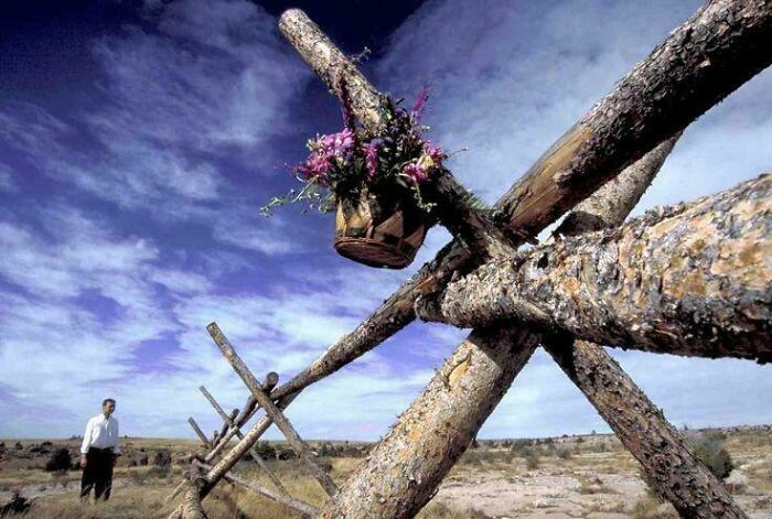 Man standing near wooden structure with flowers, under blue sky, reflecting true crime stories' eerie setting.