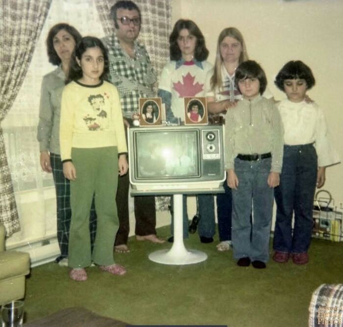 Vintage awkward family photo with seven members standing around an old TV, capturing a sweet and nostalgic moment.