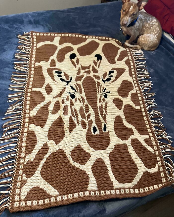 Crochet blanket with detailed giraffe pattern laid on bed next to small dog, showcasing crochet masterpiece design.