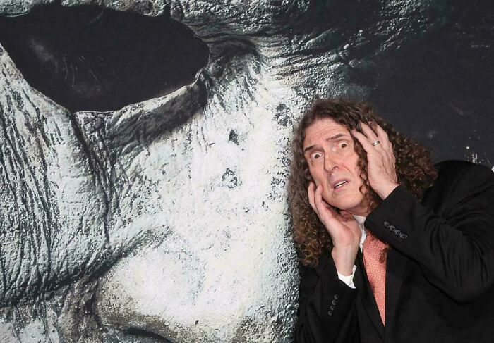 Man in a suit appears surprised next to a large, textured artwork. Fascinating scene captures attention.