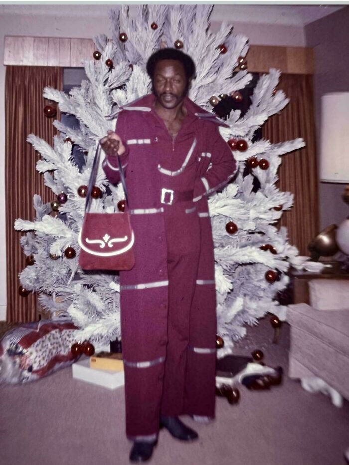 Vintage photo of a man in a maroon outfit holding a handbag by a decorated white Christmas tree, old images history class.