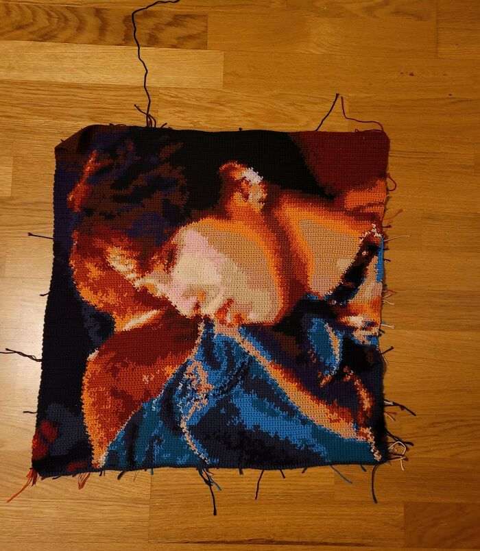 Crochet artwork depicting a detailed portrait with rich colors and intricate stitch work on a wooden floor.