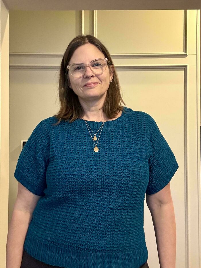 Woman wearing a teal crochet top with short sleeves and textured stitch pattern, standing indoors against a beige background.