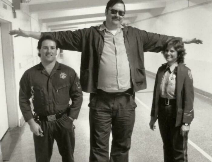 Historical photo of a tall man with arms outstretched between two officers in a hallway, showcasing people decades ago.