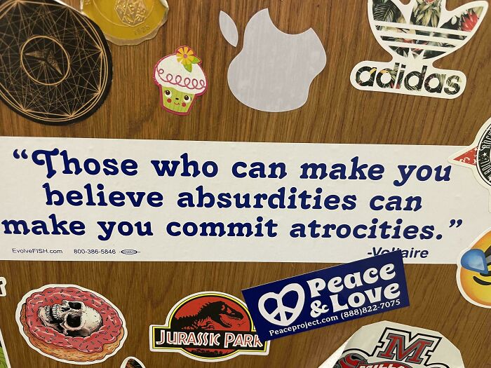 Funny bumper sticker with a Voltaire quote surrounded by various other colorful stickers on a wooden surface.