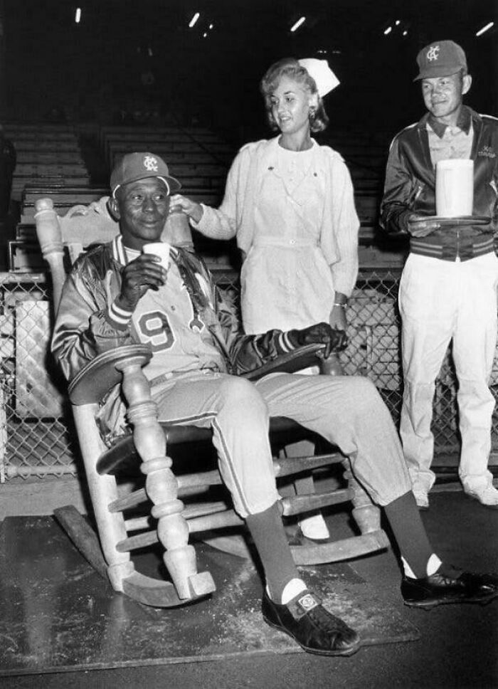 Man in a baseball uniform sitting in a rocking chair, with a nurse and another person holding a cap nearby, showcasing historical living.