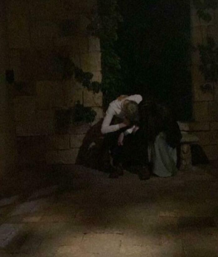 Two people sitting in a dimly lit stone courtyard, resembling a Renaissance painting scene.