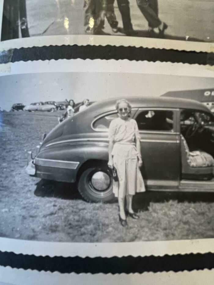 A woman standing beside a vintage car, showcasing historical fashion and everyday life from decades ago.