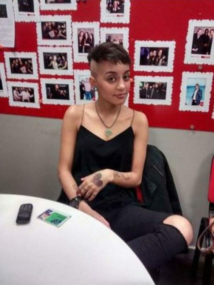 Person with short hair sitting at a table, with a photo collage on the red wall behind them, reflecting "blunder years."