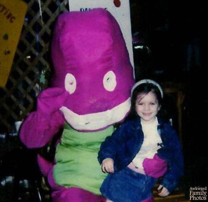 Child smiling while sitting on the lap of a person in an awkward purple dinosaur costume in a family photo setting.