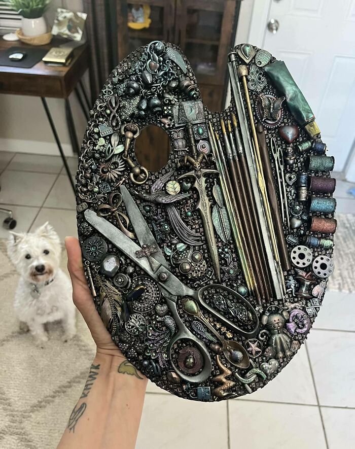 Intricate secondhand find: a detailed art palette adorned with vintage objects and tools, held above a curious small dog.