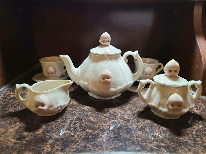 Weird secondhand tea set with baby faces, featuring a teapot, creamer, sugar bowl, and cups on a dark surface.